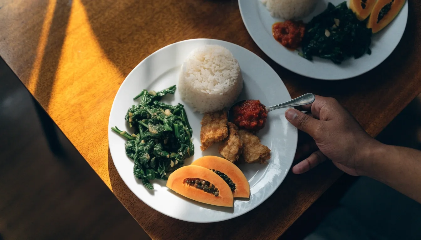 A portioned family plate of rice, greens, tempeh, sambal and papaya on a wooden table, a hand setting a second plate down