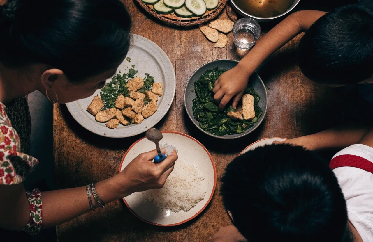 Hands lifting a bowl of stir-fried greens and tempeh from a kitchen counter set with rice and soup